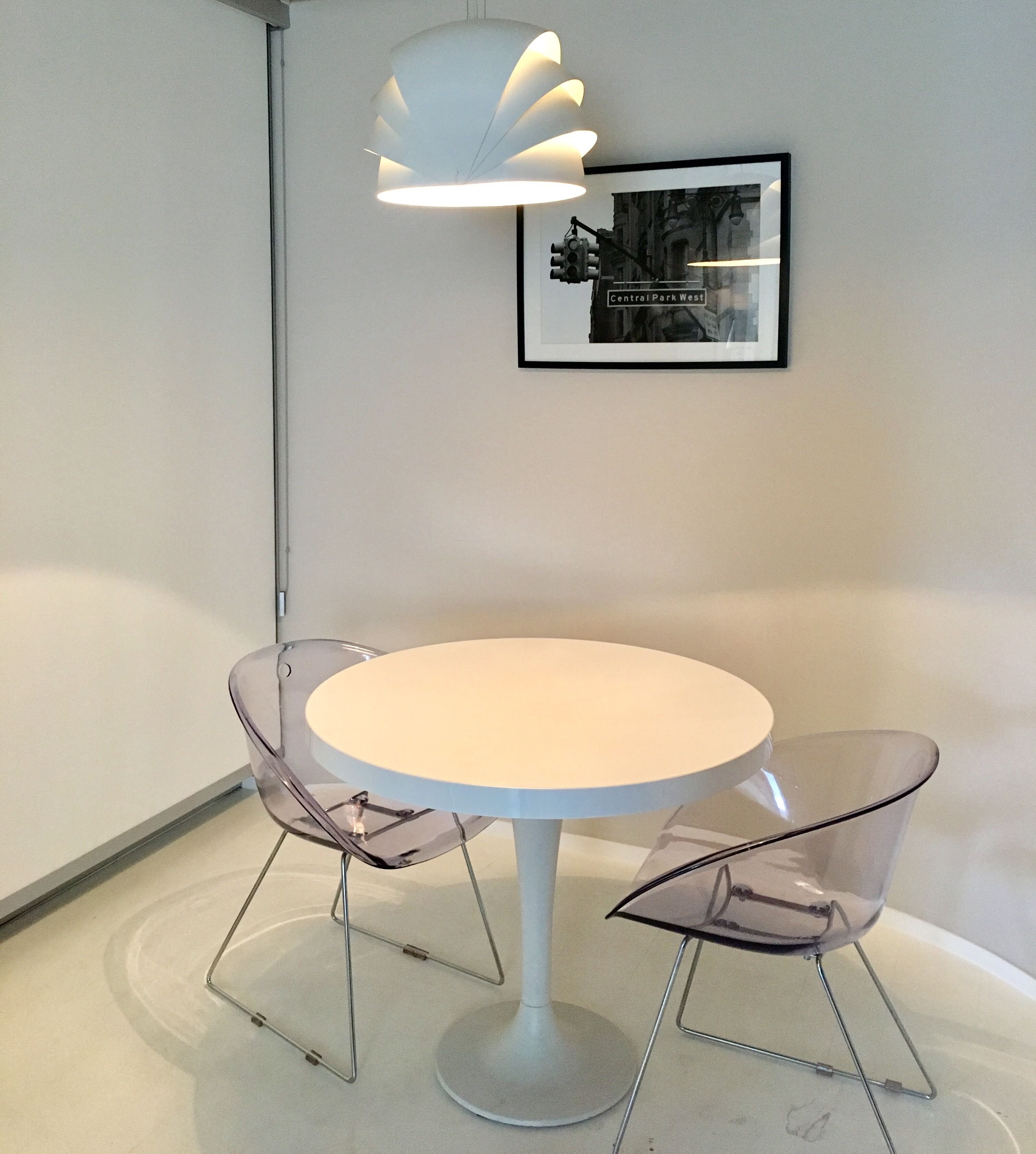 A round white table with two transparent chairs, a white pendant light, and a framed picture on the wall.