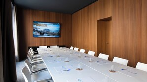 Un salón de conferencias con una larga mesa, sillas blancas y una pantalla instalada en la pared.