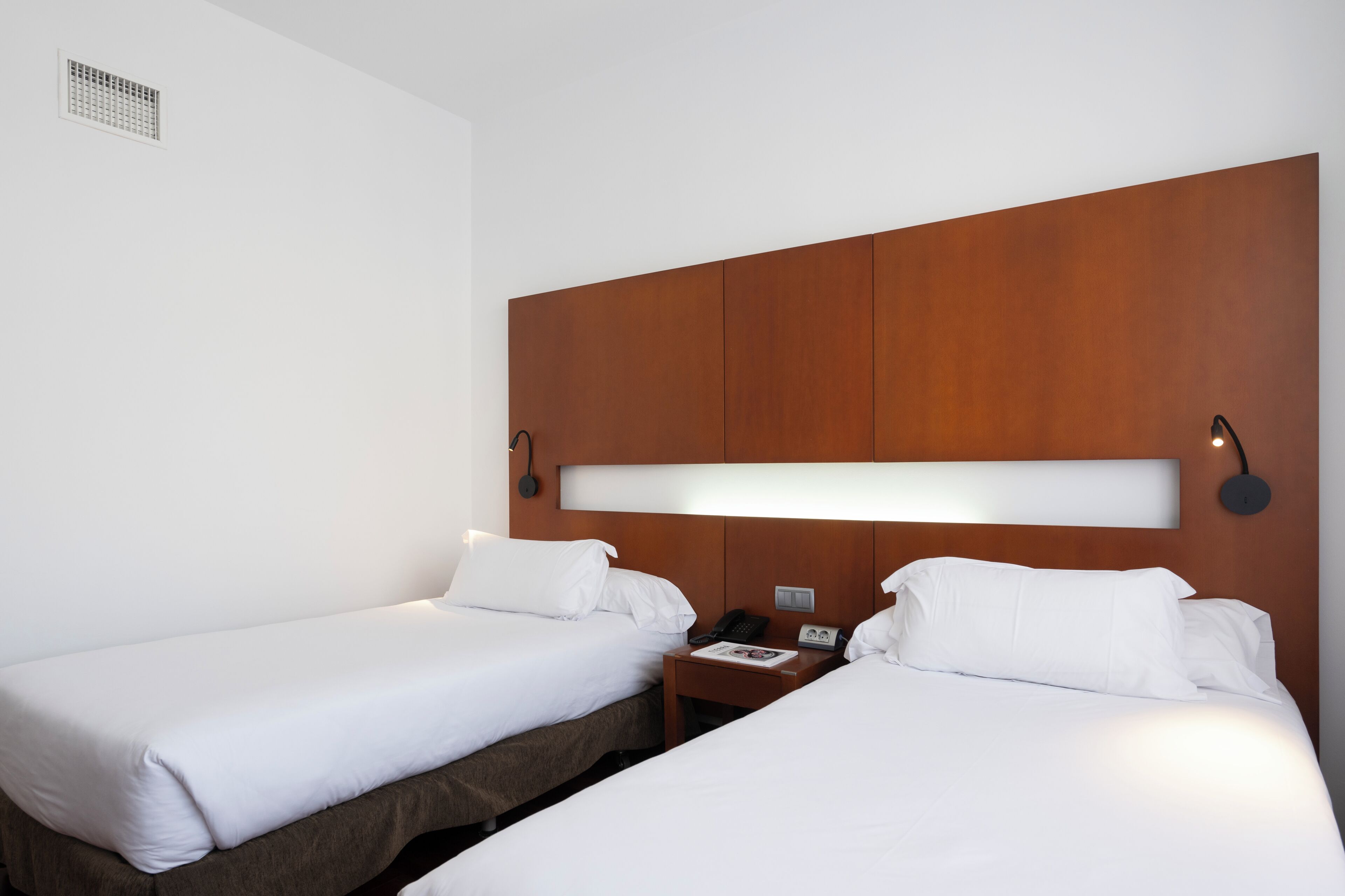 A hotel room with two single beds, a wooden headboard, a bedside table with a lamp, and a phone.