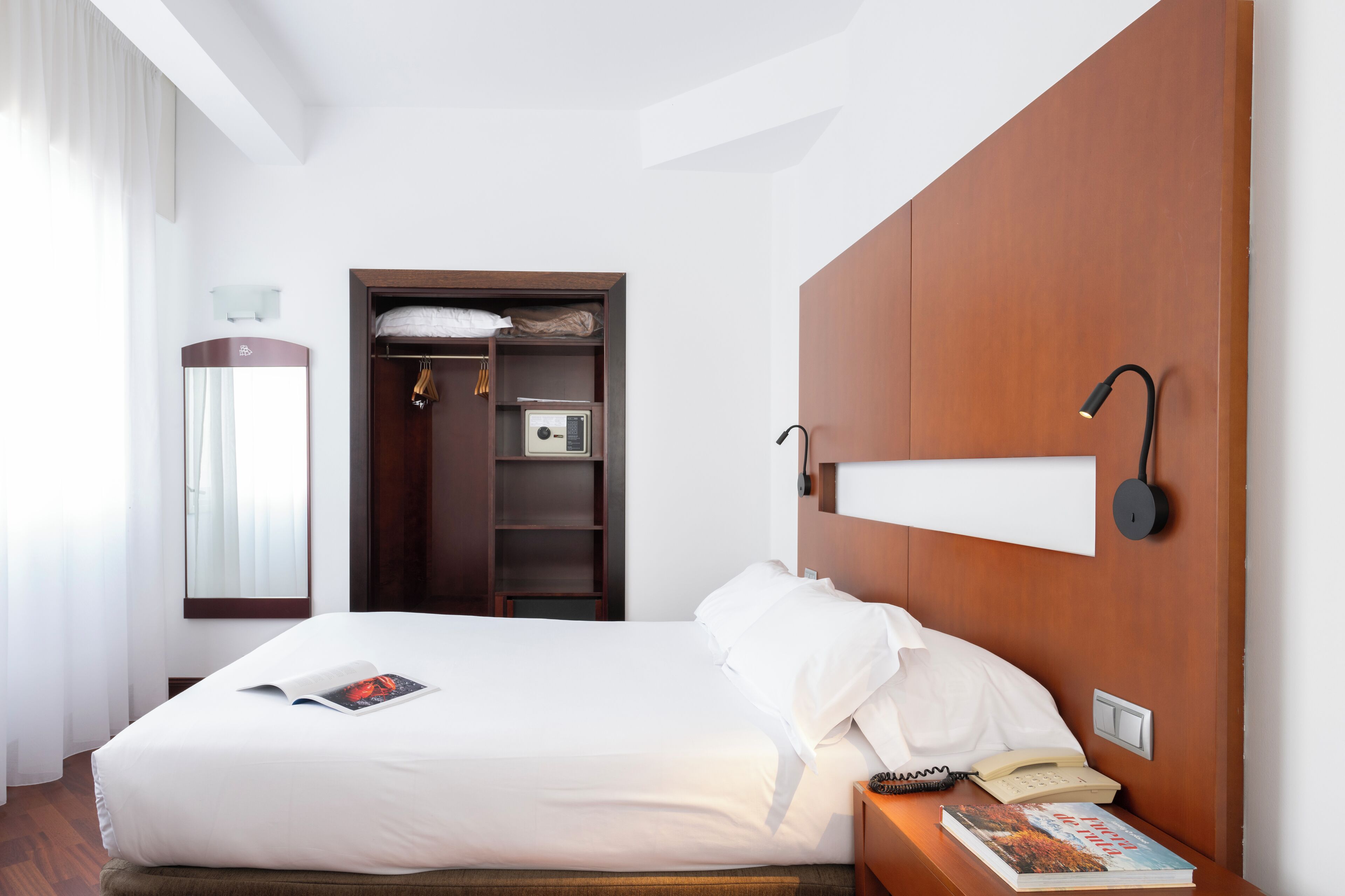 Una habitación de hotel con una cama, una mesita de noche con un libro y un teléfono, un armario, un espejo y una lámpara de pared.