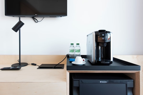 A modern hotel room with a flat-screen TV mounted on the wall, a coffee maker, two bottles of water, a remote control, and a black Dometic refrigerator.