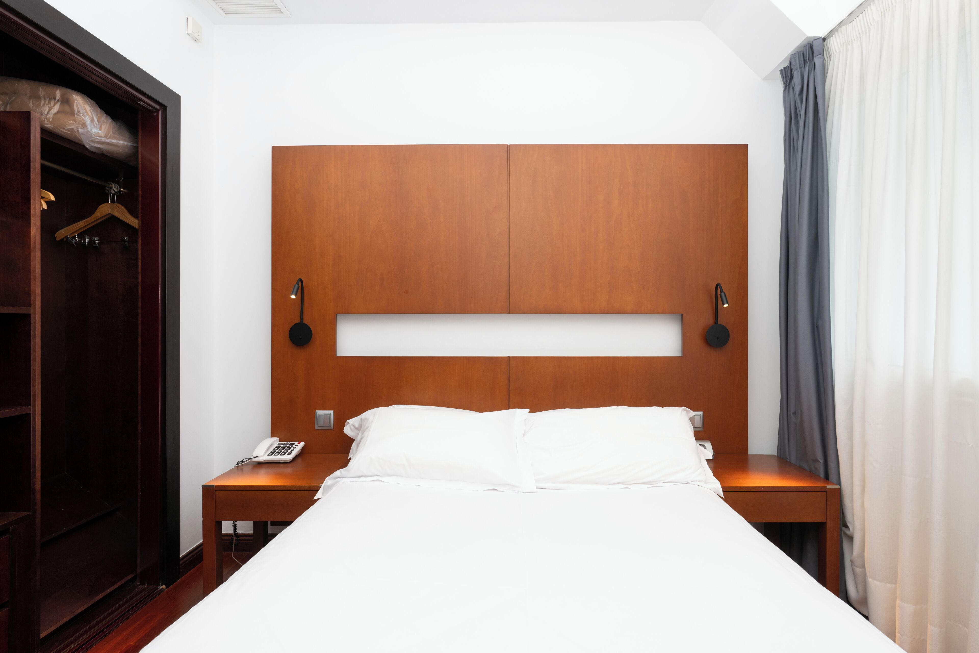 Habitació d'hotel amb un capçal de llit de fusta, un llit amb roba de llit blanca, una taula de nit amb una làmpada, un telèfon i un armari amb barra per penjar roba.