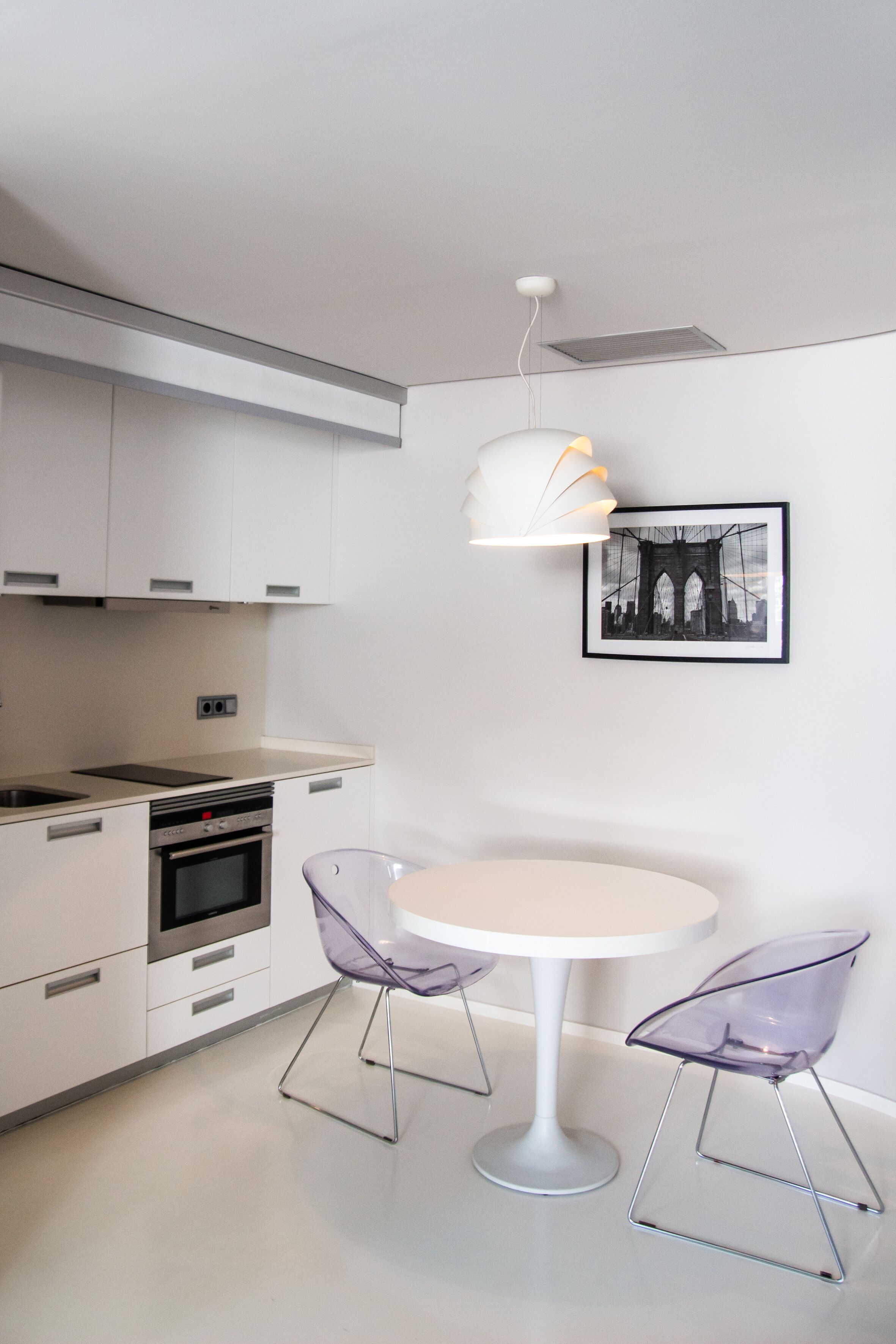 A modern kitchen with white cabinets, a built-in oven, and a round table with transparent chairs.