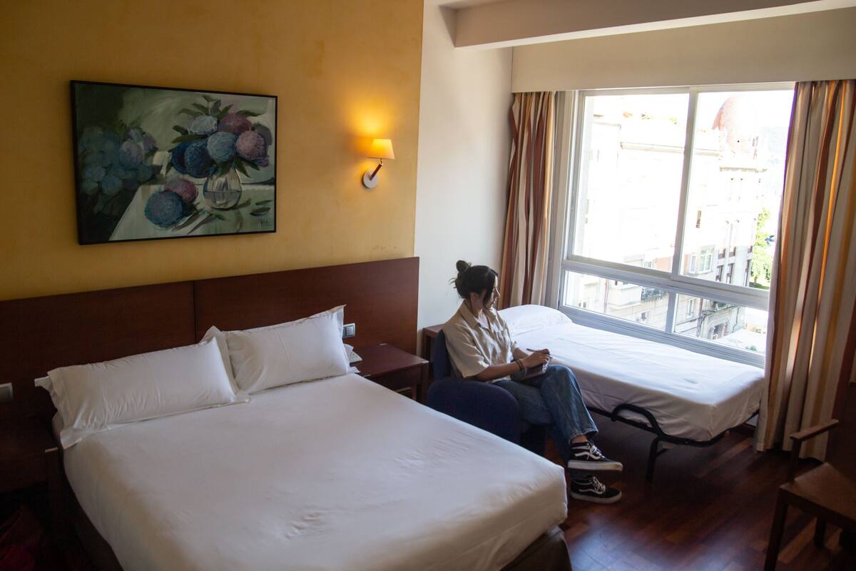 Una habitación de hotel con dos camas, un cuadro en la pared, una ventana con cortinas y una mesita de noche.