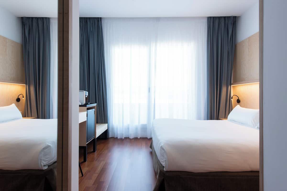 Habitación de hotel con dos camas, un escritorio y un ventanal con cortinas.