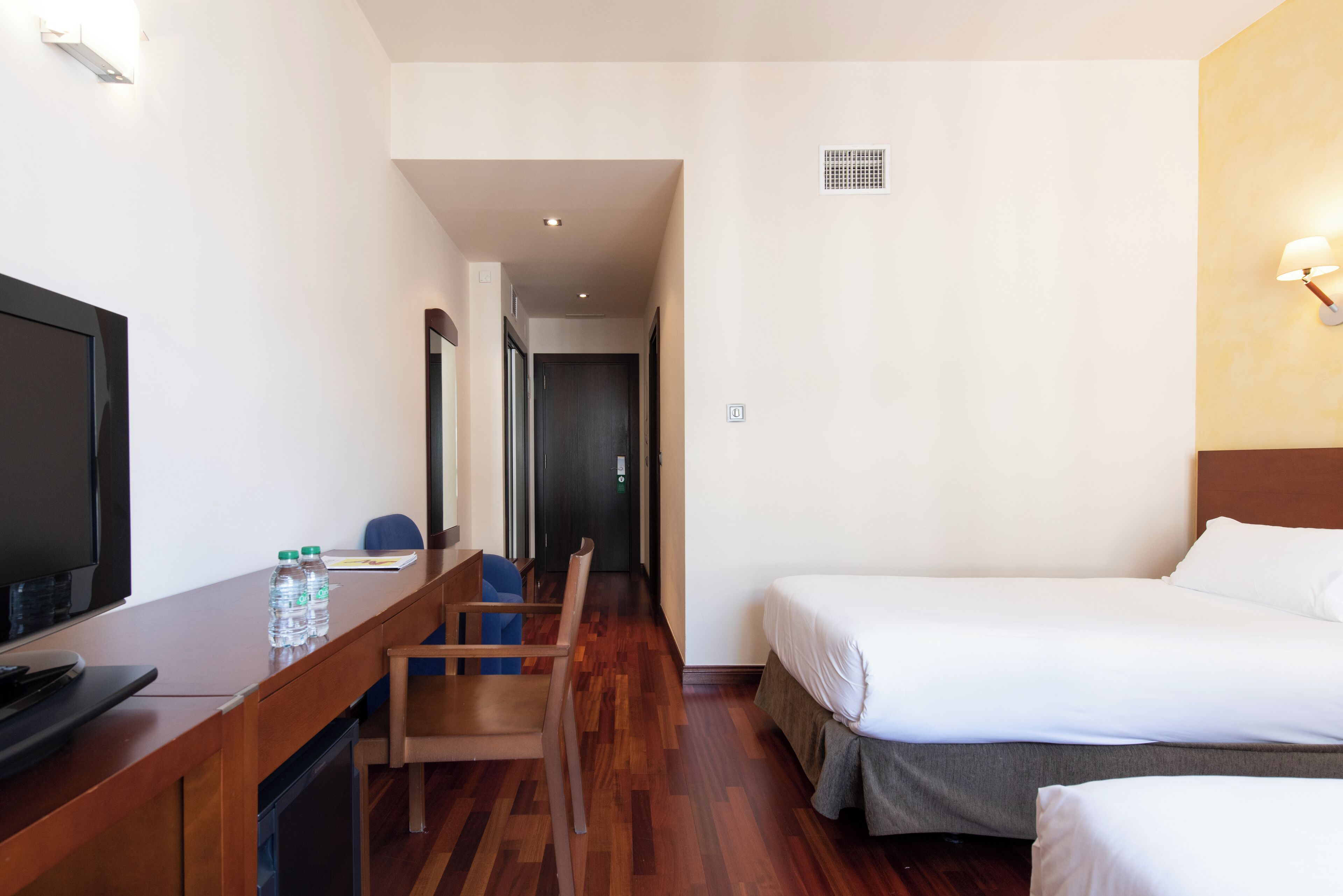A hotel room with a bed, a desk with a television, a chair, and a water bottle.