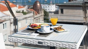 Una mesa de desayuno con un vaso de jugo de naranja, una taza de café y un plato de fruta y pastelería en un balcón con vista a un paisaje urbano.