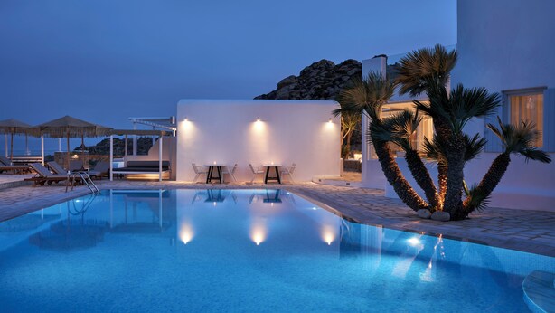 Una piscina con sillas de descanso y mesas, rodeada de palmeras y un edificio de fachada blanca.