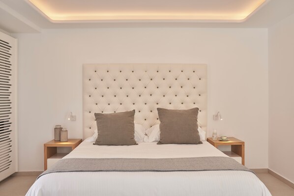 Un dormitorio moderno con cabecera capitonada, una cama grande con dos almohadas, una mesita de noche con lámpara y un tocador con un florero.