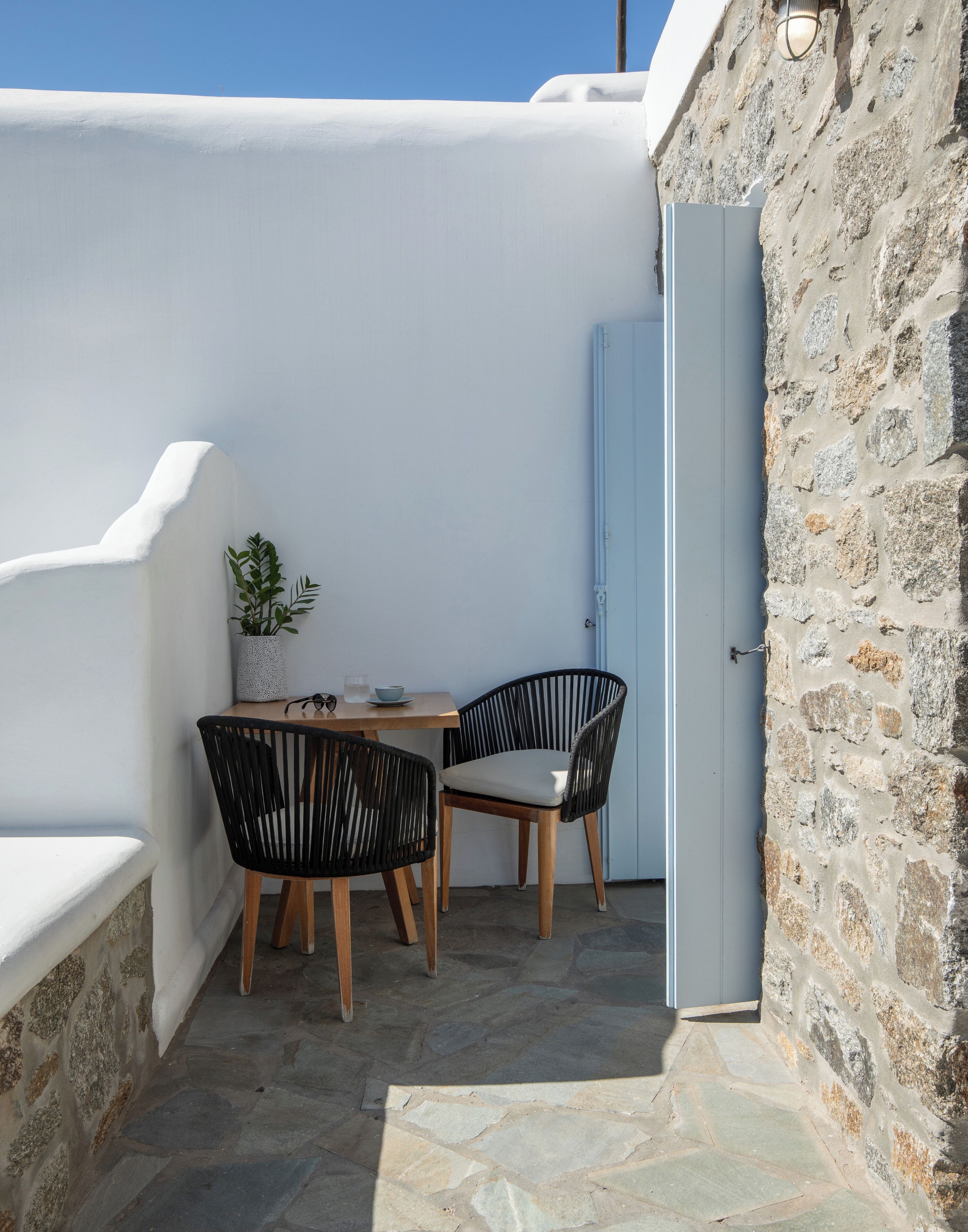 A small outdoor seating area with two black chairs and a wooden table, a potted plant, and a stone wall.
