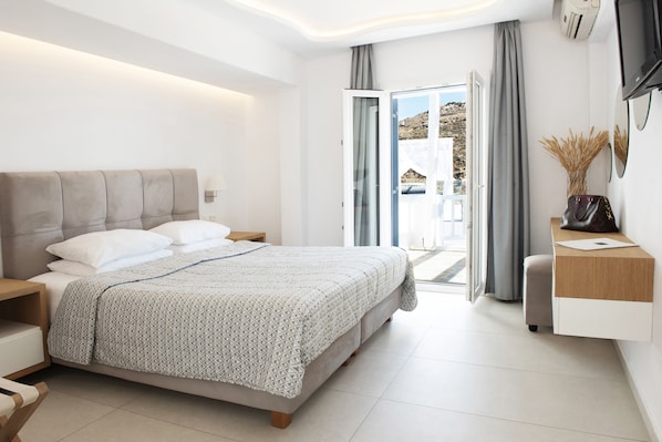 A modern bedroom with a large bed, a bedside table, a nightstand, a desk with a chair, and a view of the outdoors through a glass door. - Paradise View Hotel (Mykonos)