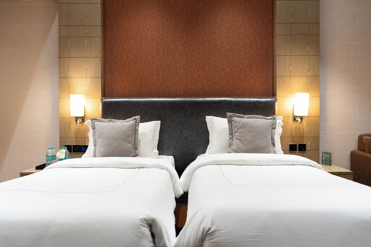 Habitación de hotel con dos camas, cabecero de madera, lámparas fijadas a la pared, mesita de noche con una botella de agua y una pequeña mesa con una silla.