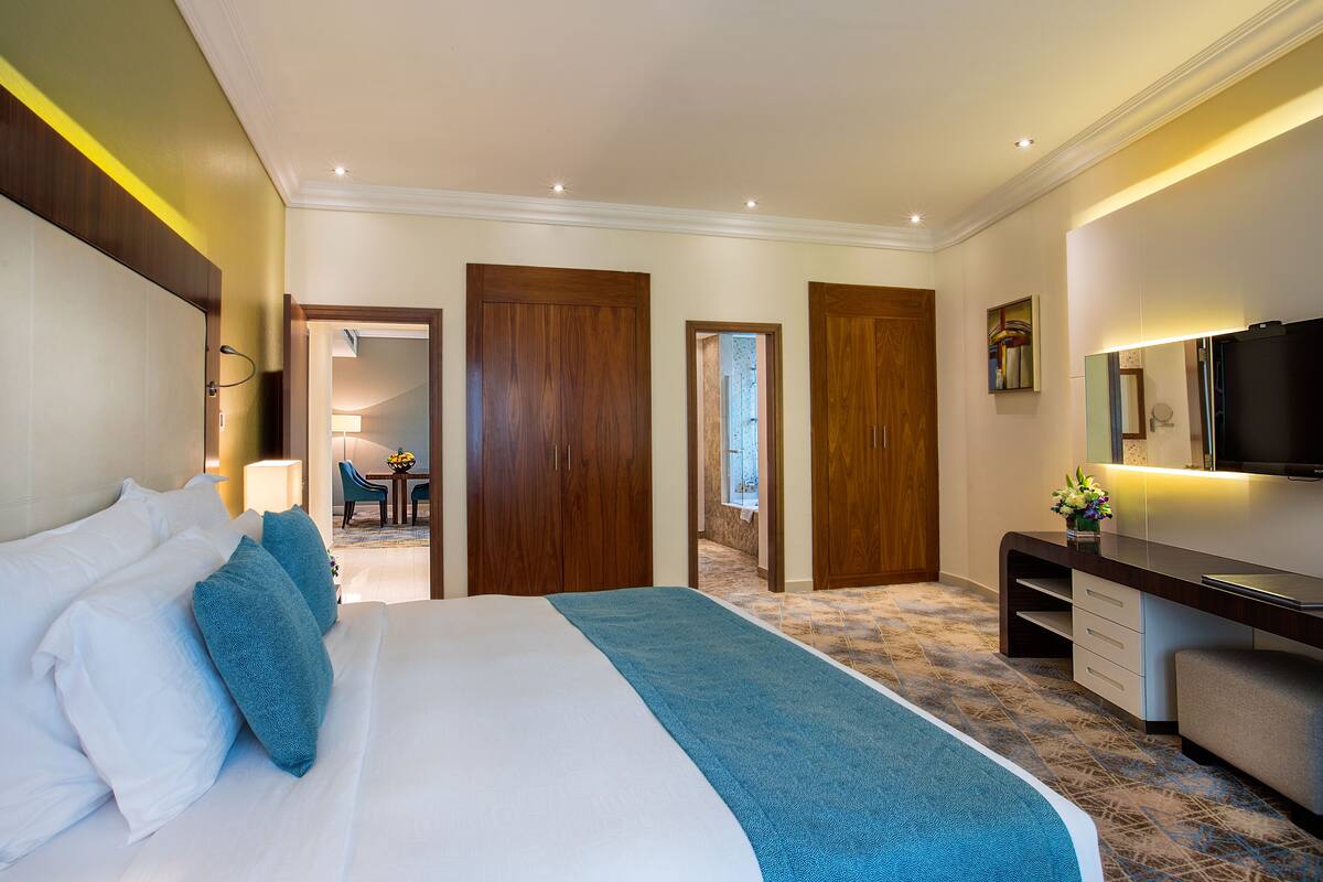A hotel room with a large bed, a desk, and a TV. There are wooden doors and a view of the hallway.