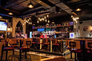 A bar with multiple stools, a bartender, and a well-stocked shelf with various bottles.
