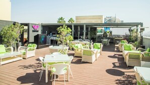 Un patio al aire libre con mobiliario de mimbre, mesas y sillas, y un edificio con un cartel al fondo.