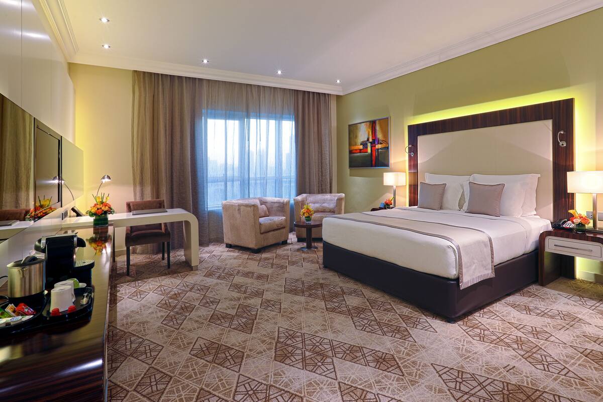 A hotel room with a large bed, a desk with a coffee maker, two armchairs, and a large window with curtains.