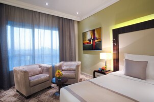 A hotel room with a large bed, two armchairs, a small table with a vase of flowers, a painting on the wall, and a window with curtains.