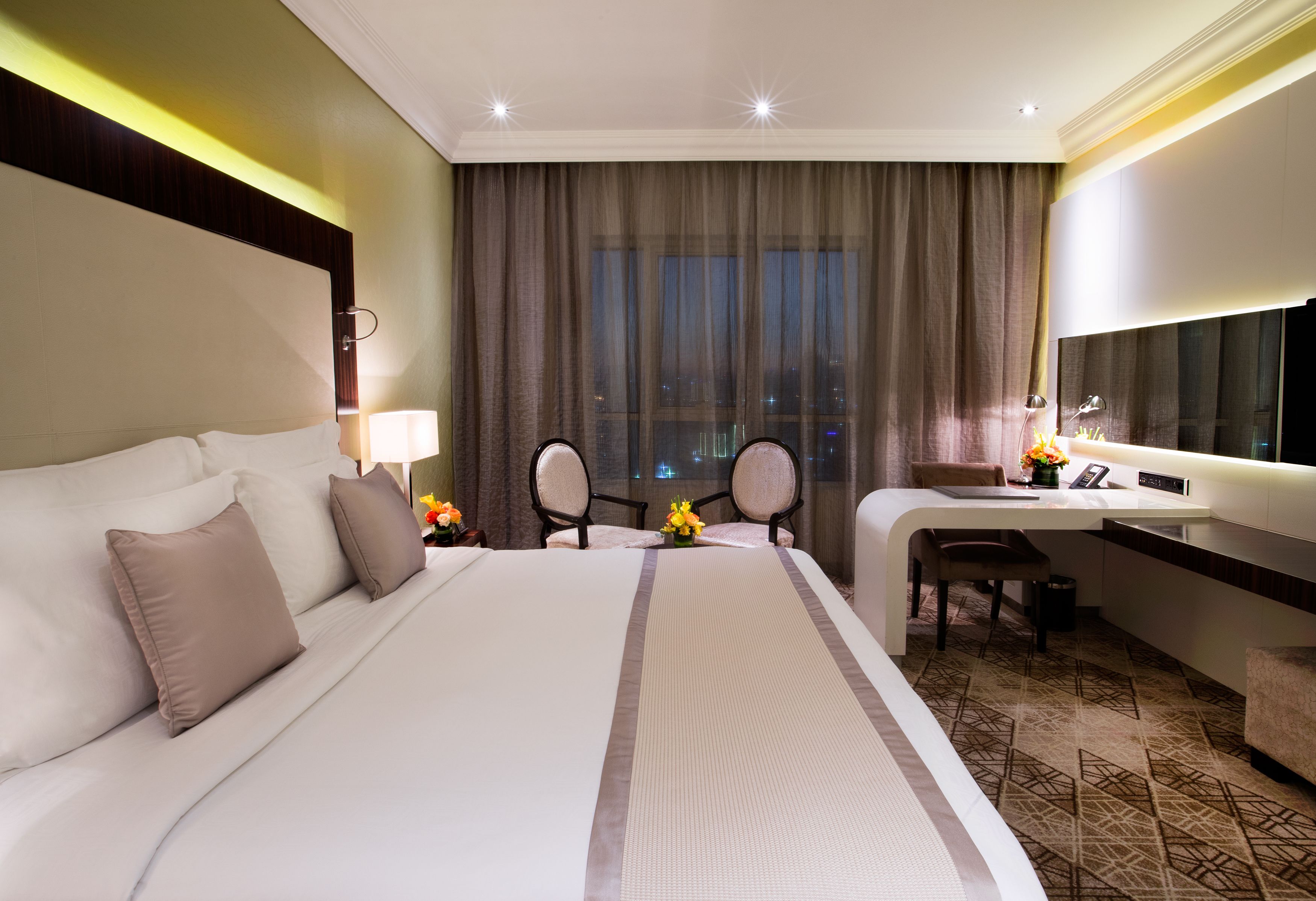 A hotel room with a large bed, a desk, and a chair. There is a window with curtains, a lamp, and a small table with flowers.