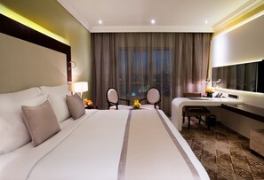 A hotel room with a large bed, a desk, and a chair. There is a window with curtains, a lamp, and a small table with flowers. - Elite Byblos Hotel (Dubai)