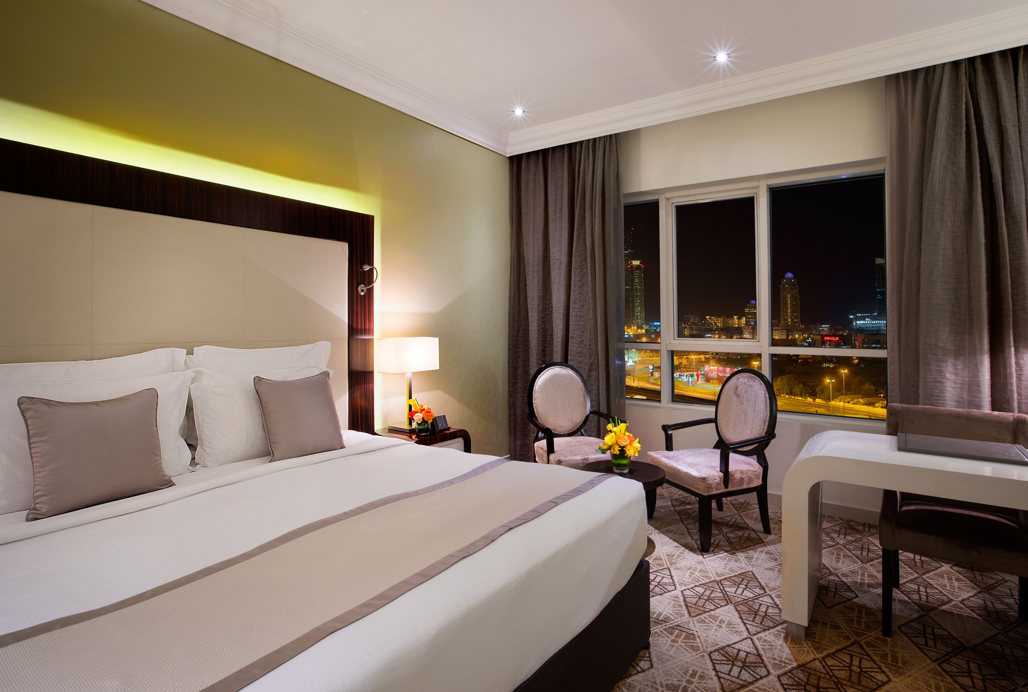 Habitación de hotel con una cama grande, dos sillas, una mesita y vistas a la ciudad de noche.