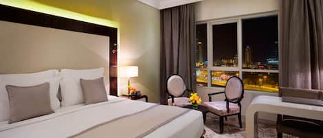 Habitación de hotel con una cama grande, dos sillas, una mesita y vistas a la ciudad de noche.