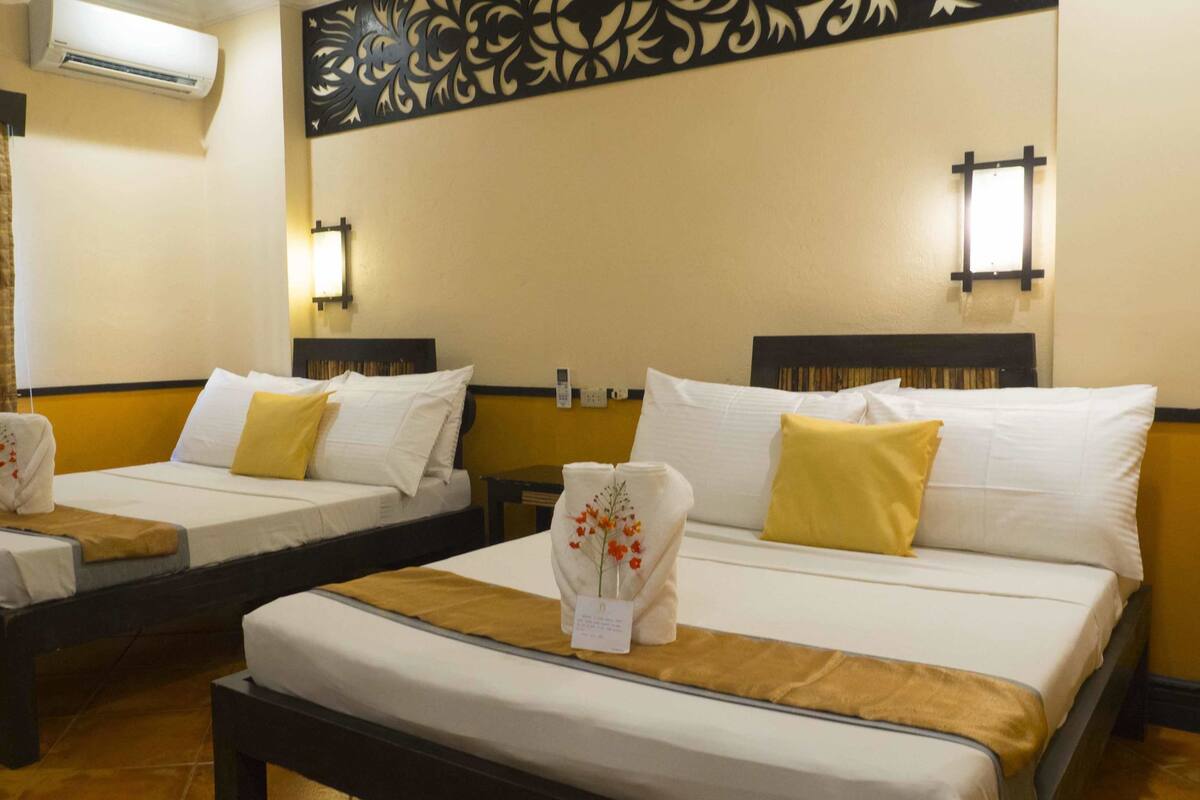 Habitación de hotel con dos camas individuales, cada una con cabecero de madera, ropa de cama blanca y un cojín decorativo amarillo. Se observa un aire acondicionado de pared y un panel decorativo sobre las camas.