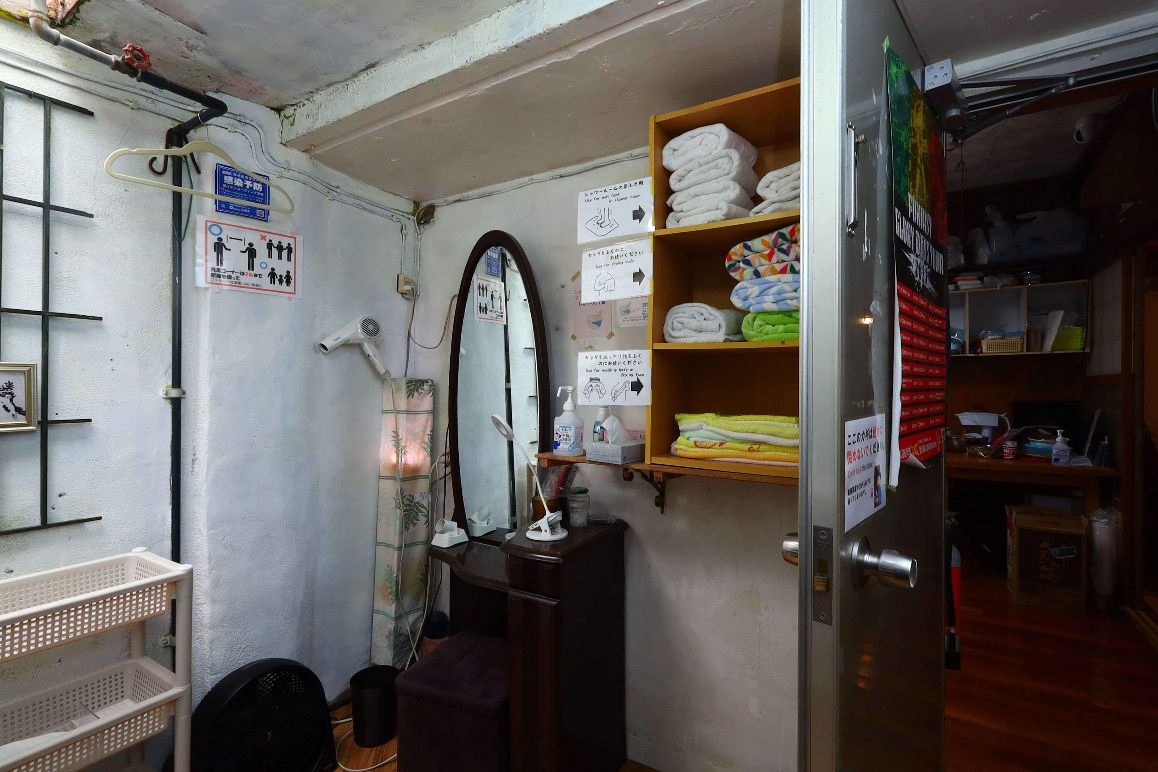 A small, compact room with a mirror, a fan, and a shelf with folded towels. There are signs on the wall and a door with a handle.