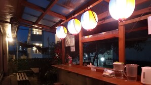 Un área de bar al aire libre con taburetes de madera, una barra con distintos objetos y faroles de papel.