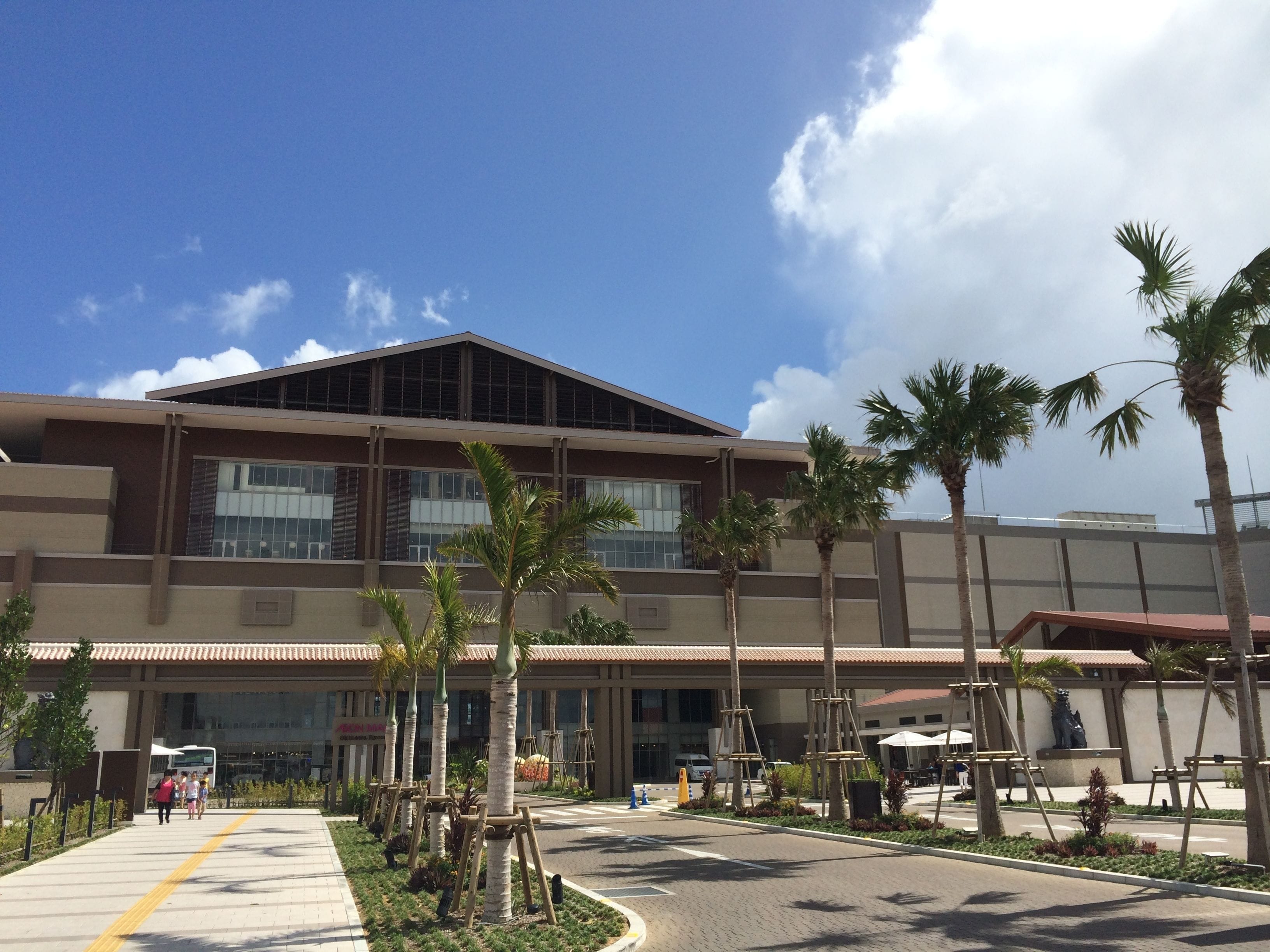 A modern building with a flat roof and large windows, surrounded by palm trees and a paved area with a yellow line marking.