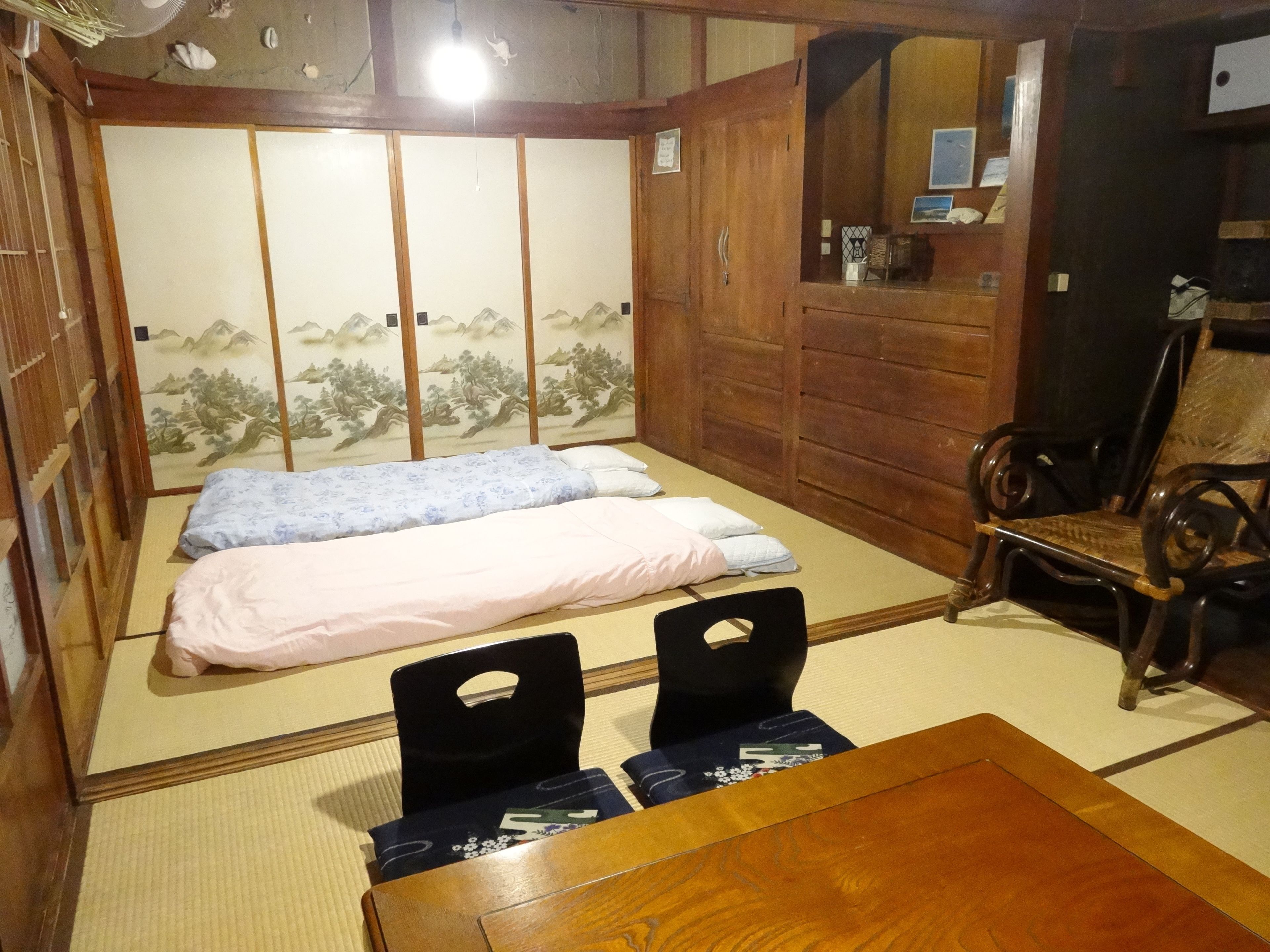 Una habitación japonesa tradicional con suelo de tatami, una cama de madera baja con dos almohadas, una silla negra y una mesa de madera.