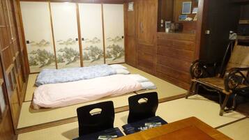 Una habitación japonesa tradicional con suelo de tatami, una cama de madera baja con dos almohadas, una silla negra y una mesa de madera.