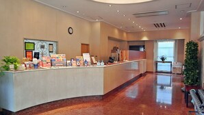 A reception area with a counter displaying informational brochures, a clock on the wall, and a large potted plant to the right. - Hotel Kizankan (Tokyo)