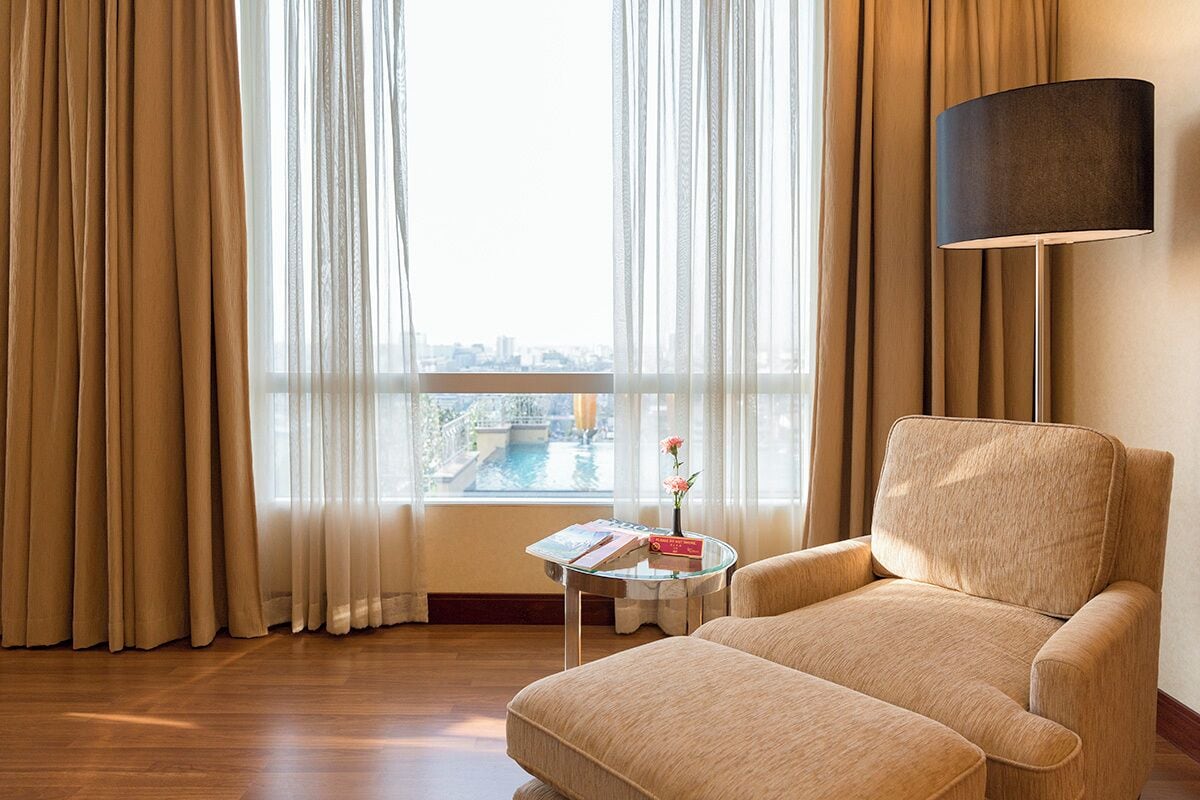 Habitación de hotel con un ventanal grande, un sofá, una mesita auxiliar con un libro y un jarrón, y una lámpara de pie.