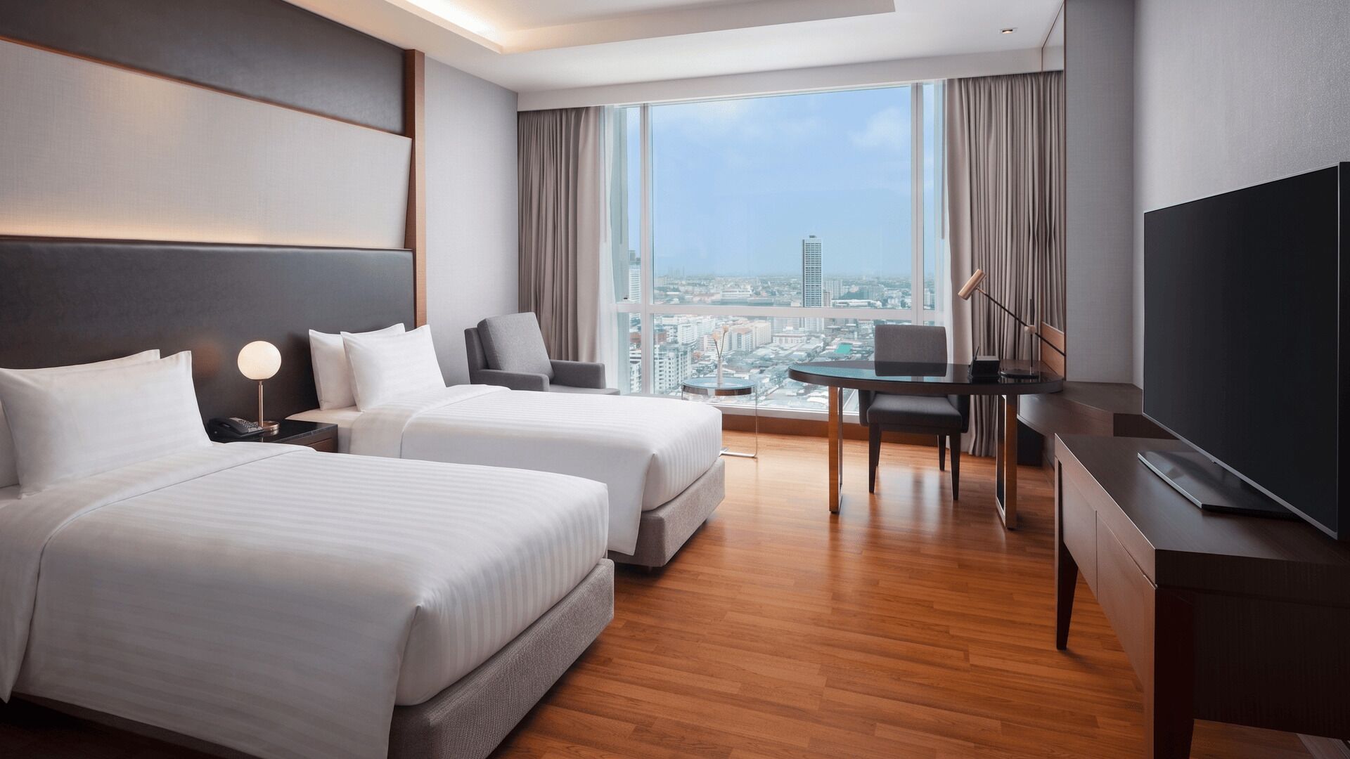 Habitación de hotel con dos camas, un escritorio, una silla, un televisor y un ventanal con vistas a la ciudad.