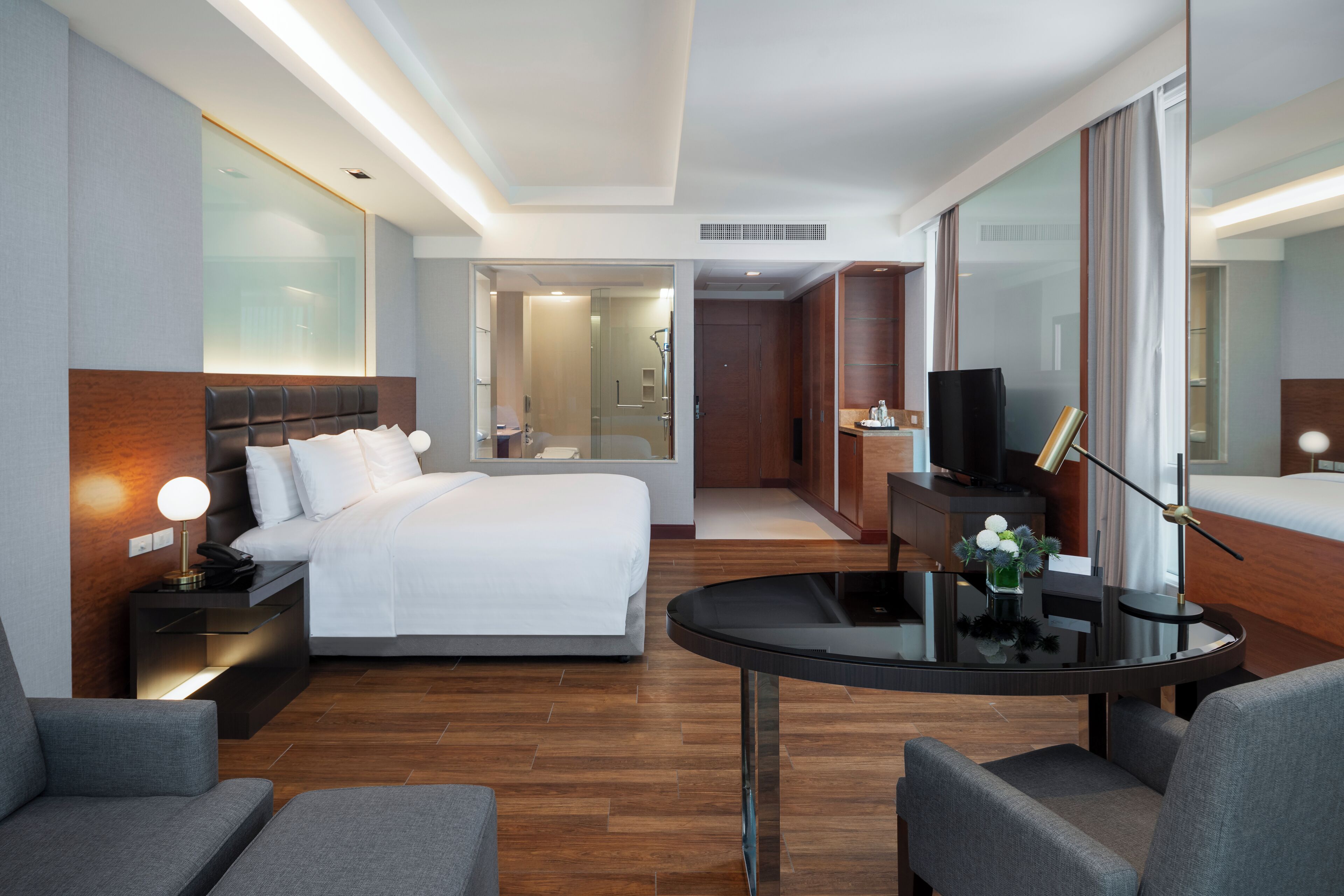 Una habitación de hotel moderna con cama, mesita de noche, lámpara, televisor y una pequeña mesa redonda con un florero.