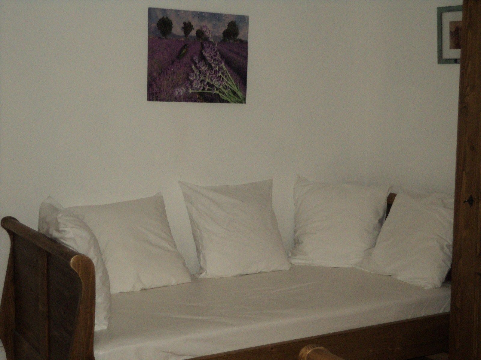 A room with a wooden bed frame, white bedding, and a painting of a field with trees on the wall.