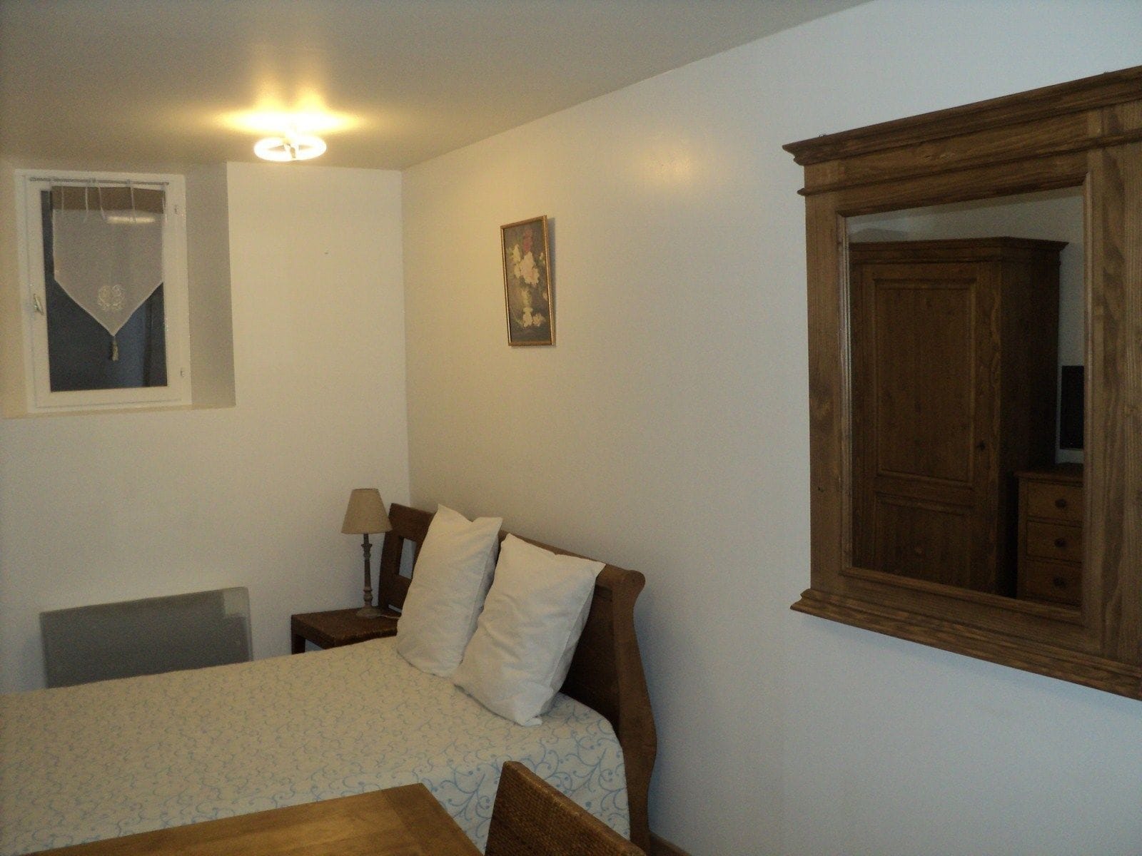 Een slaapkamer met een bed, een nachtkastje, een lamp, een houten garderobe, een raam met gordijn en een lijst met een foto aan de muur.