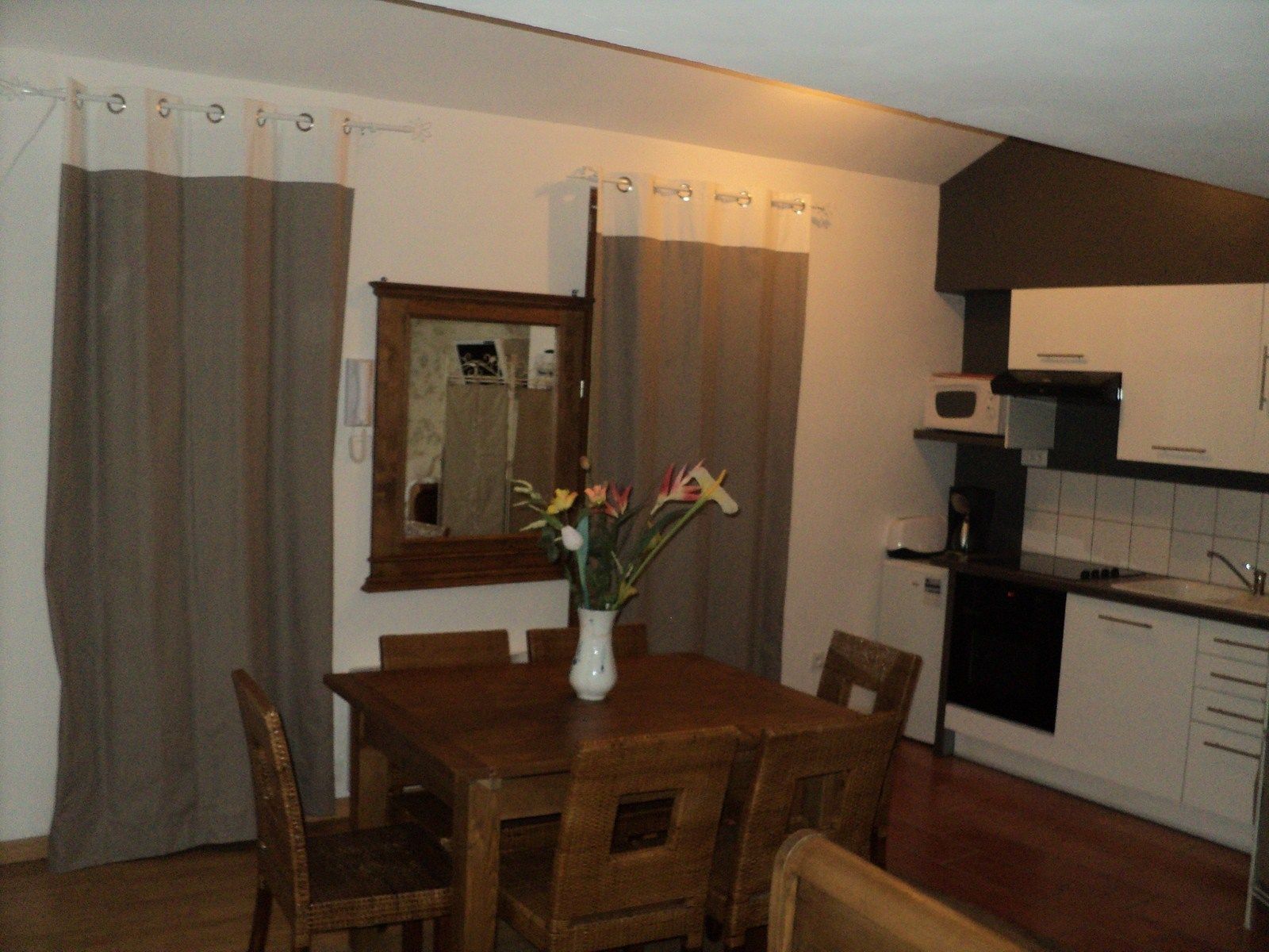 A dining area with a wooden table and chairs, a vase with flowers, and a kitchen with white cabinets and a microwave.