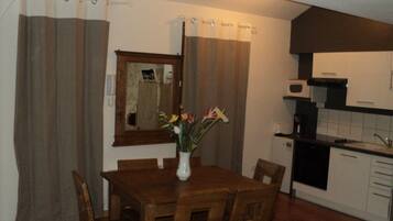 A dining area with a wooden table and chairs, a vase with flowers, and a kitchen with white cabinets and a microwave.