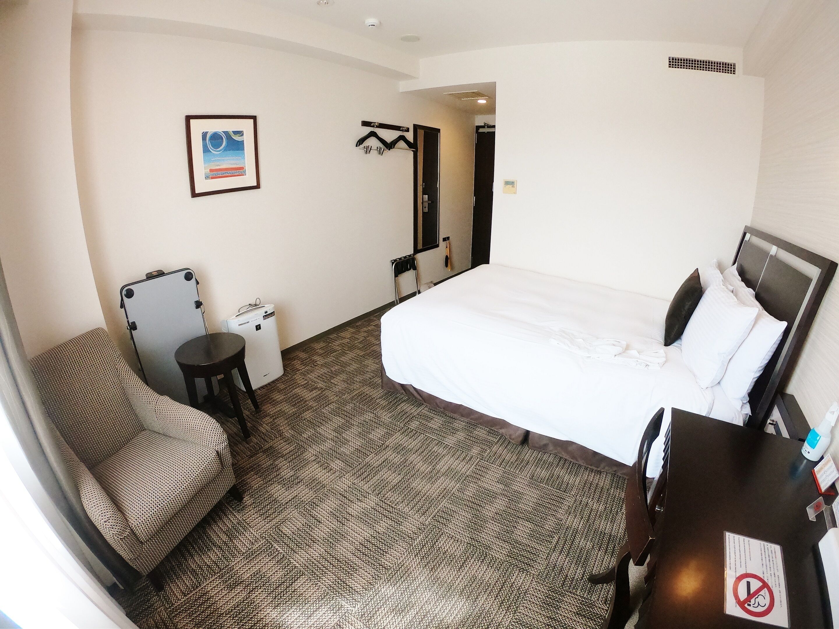 Une chambre d’hôtel avec un grand lit, une chaise, une petite table, un porte-manteau et un tableau au mur.