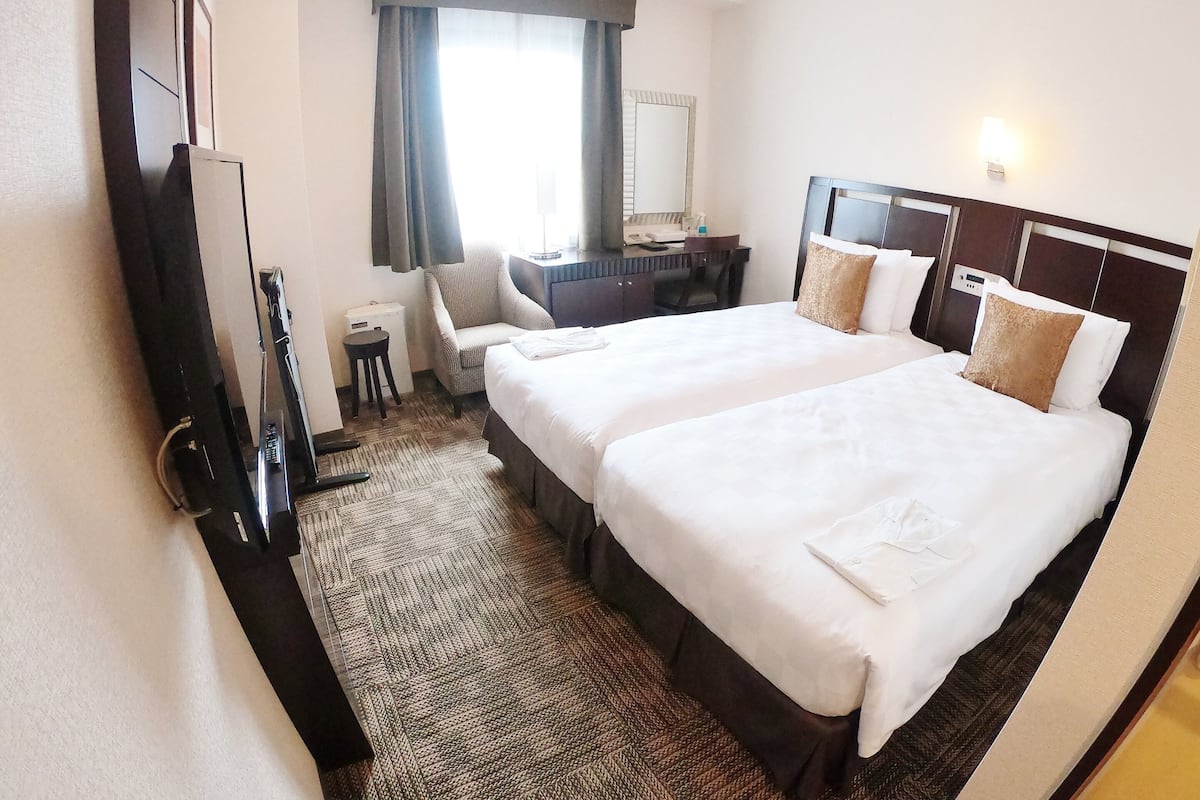 A hotel room with two beds, a desk, a chair, a TV, and a window with curtains.