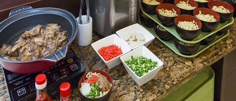 A kitchen counter with a pot of meat, a rice cooker, and bowls of ingredients.