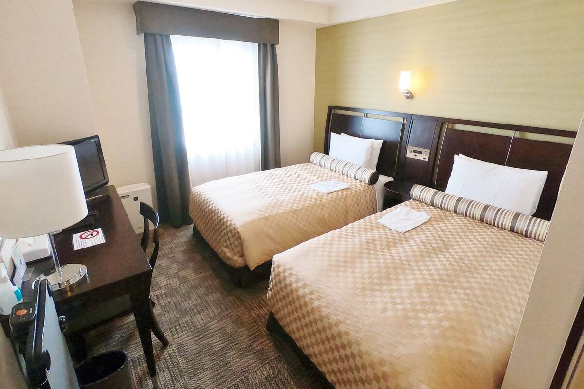 A hotel room with two beds, a desk with a computer, a lamp, and a window with curtains.