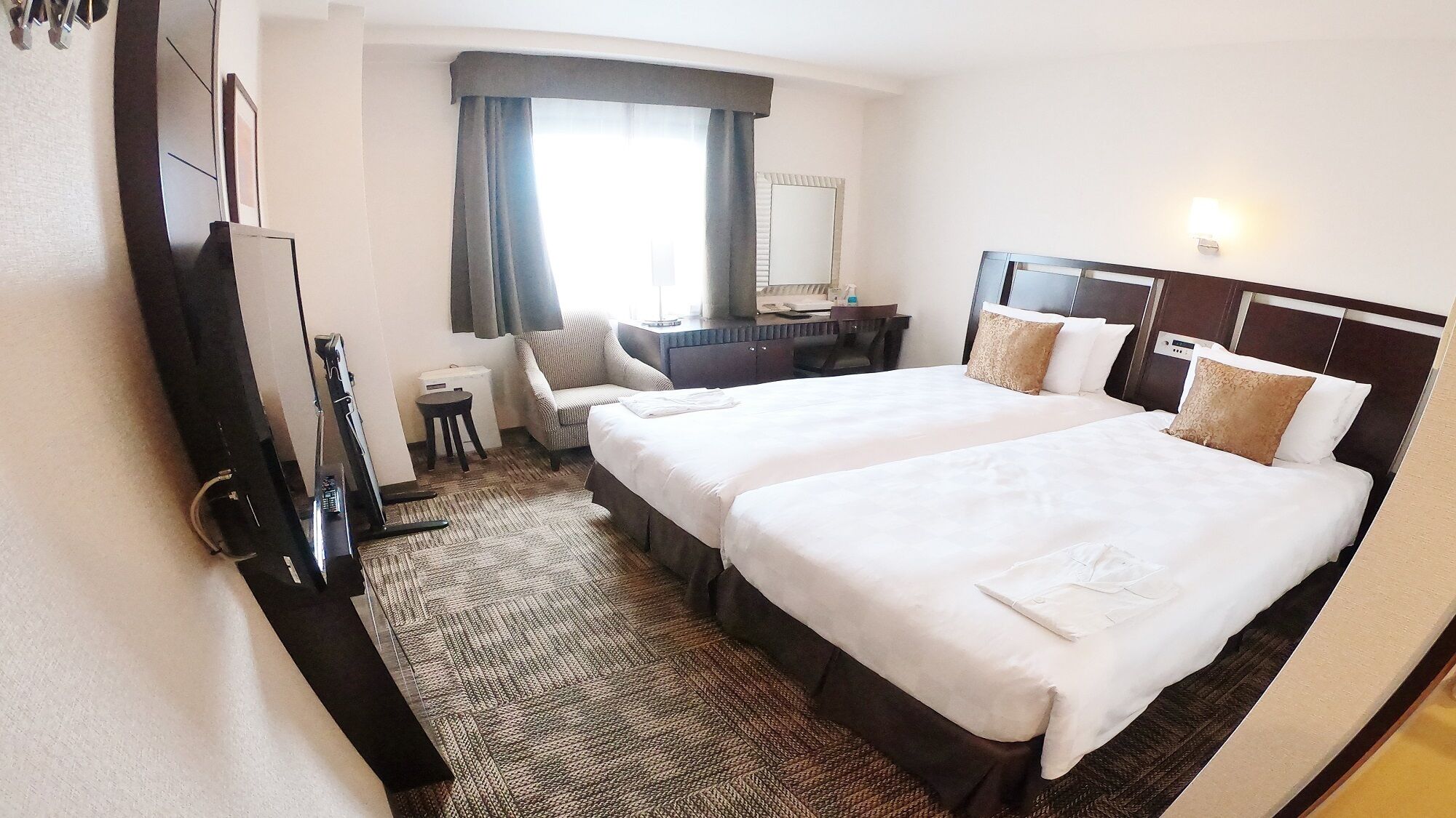 Une chambre d’hôtel avec deux lits, un bureau, une chaise, une petite table, un miroir et une fenêtre avec des rideaux.