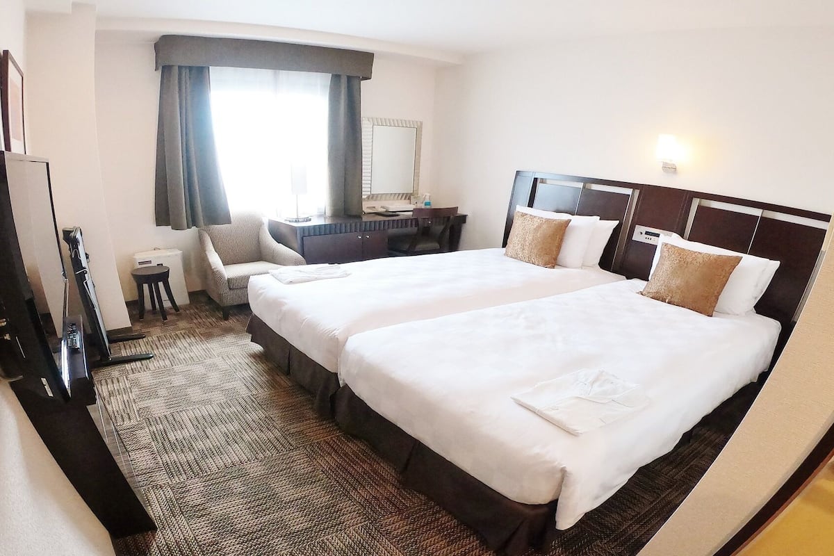 A hotel room with two beds, a desk, a chair, a small table, a mirror, and a window with curtains.