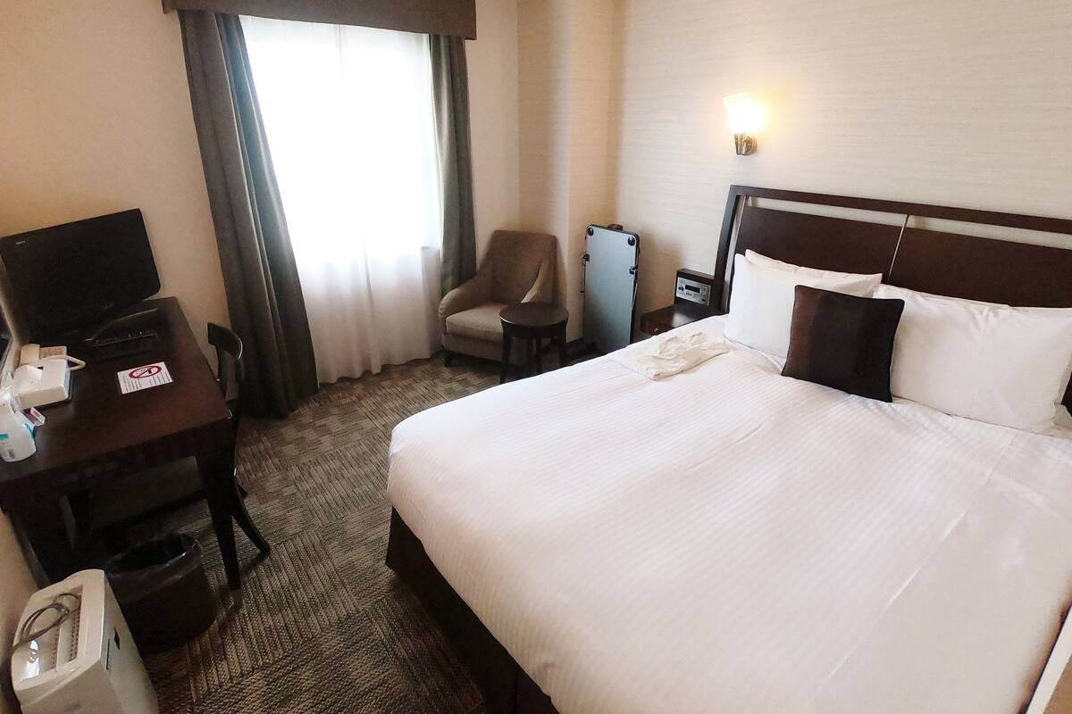 A hotel room with a bed, a desk with a computer, a chair, a nightstand, and a lamp.