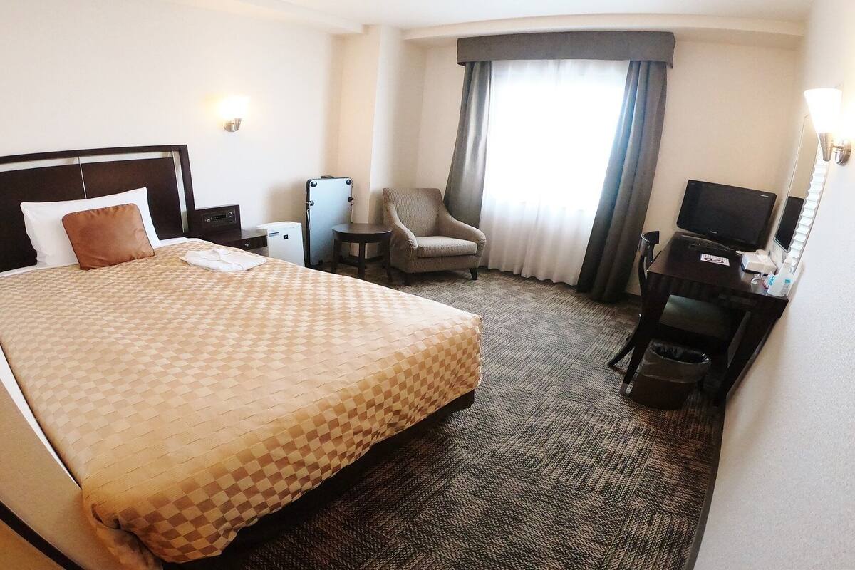 A hotel room with a large bed, a chair, a desk with a television, and a small table with a lamp.