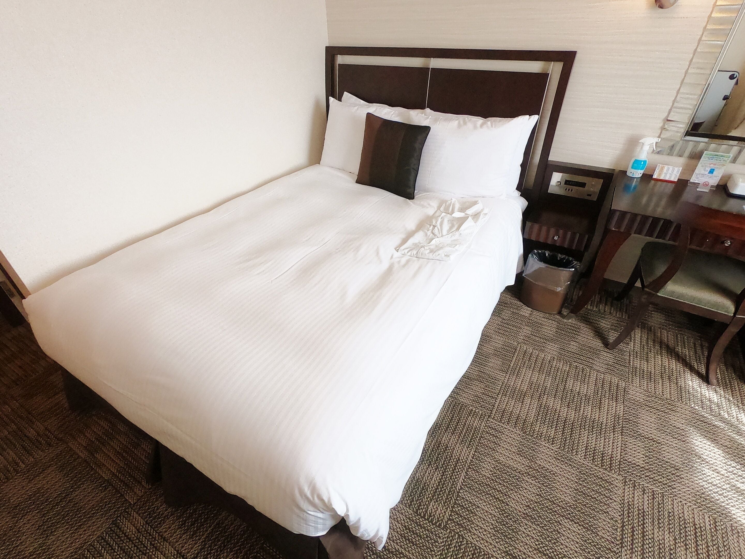 A neatly made bed with white linens, a wooden headboard, and a small bedside table with a bottle and a tissue box.