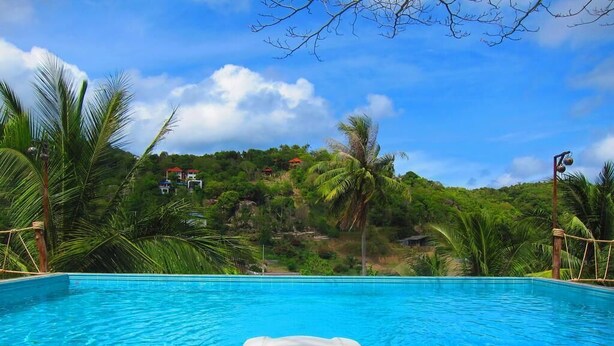 A swimming pool with a lounge chair in the foreground, surrounded by lush greenery and a hill with buildings in the background. - Koh Tao Toscana (Koh Tao)