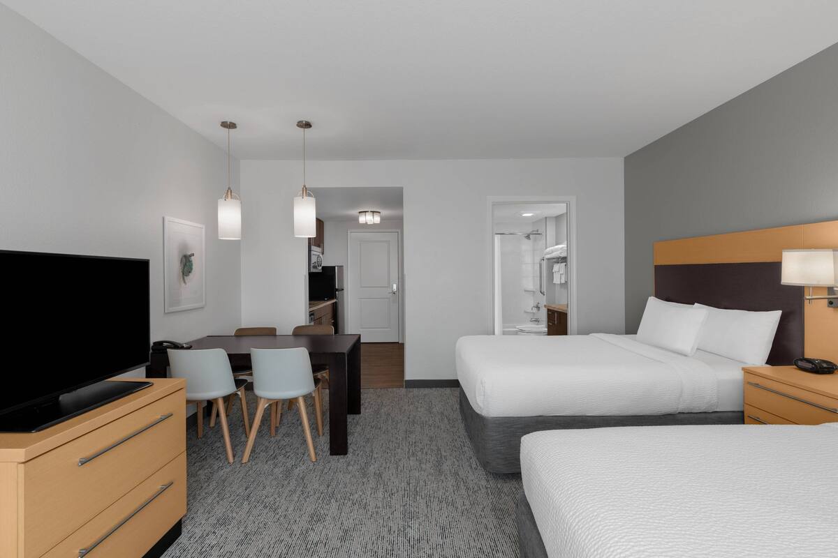 Habitación de hotel con cama, mesita de noche, televisión, escritorio, sillas y piso alfombrado.